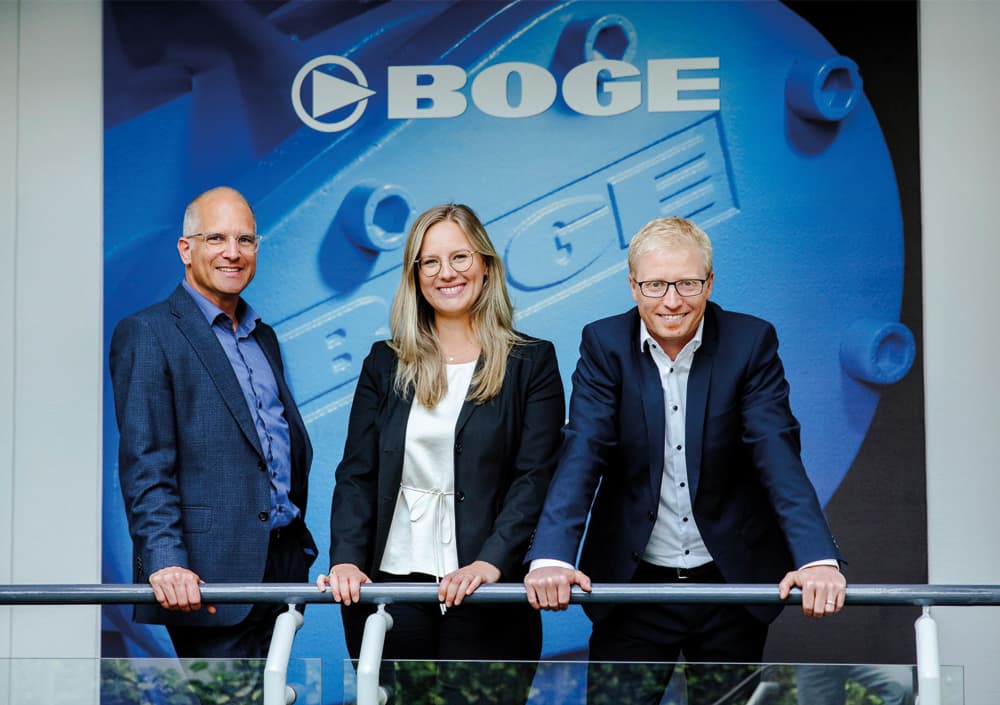 Von links nach rechts: Dr. Sebastian Göbel (Geschäftsführer), Julia Meier-Scheuven (Business Development Managerin, fünfte Inhabergeneration) und Dr. Björn Six (Geschäftsführer) vor dem BOGE Logo am Unternehmensstandort Bielefeld.