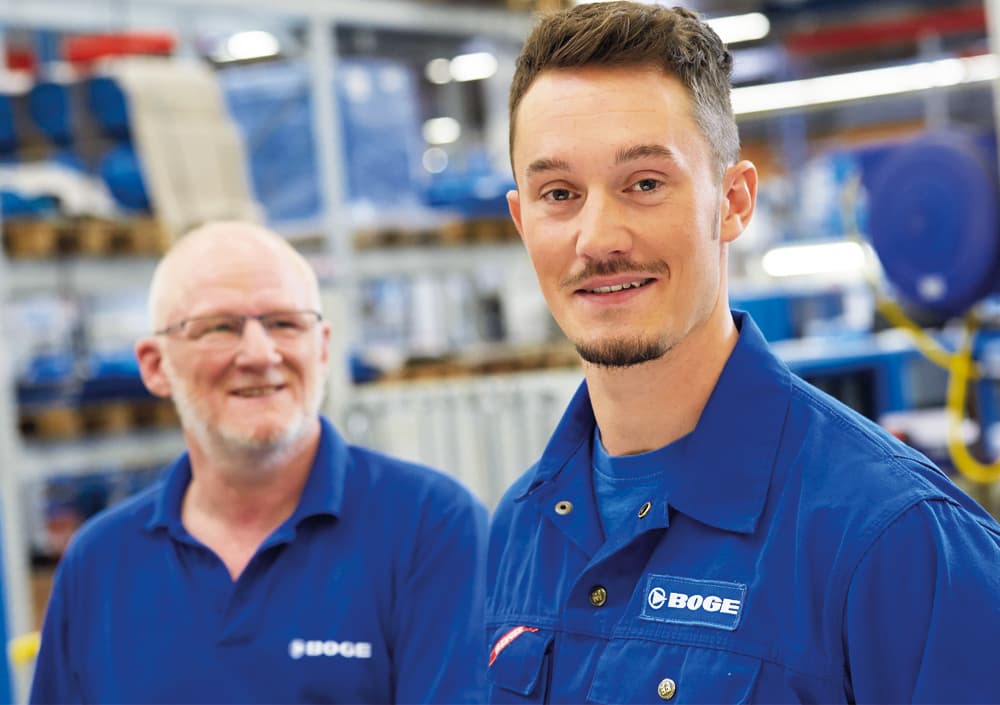 BOGE Mitarbeiter stehen zu zweit nebeneinander mit blauer BOGE Kleidung, ein jünger und ein älterer Mitarbeiter