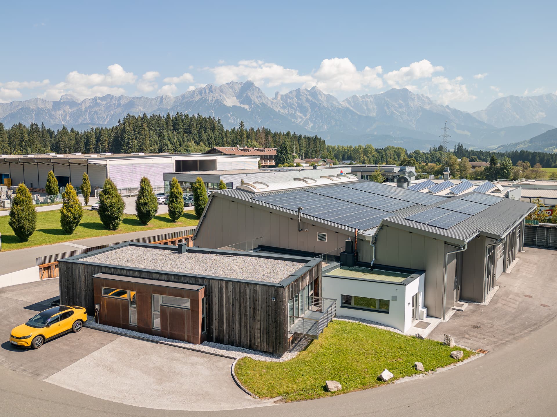 Modern industrial building with solar panels on the roof, surrounded by greenery and mountains in the background, with a yellow car parked outside.