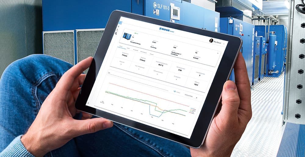 Mann haellt Remote Monitoring Screen auf Tablet vor Container mit Kompressoren