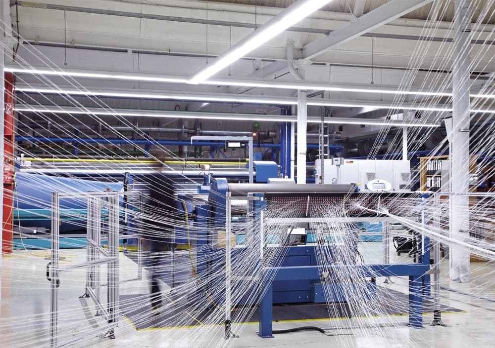 Eine blaue Webmaschine mit Fäden in alle Richtungen in einer beleuchteten Fabrikhalle