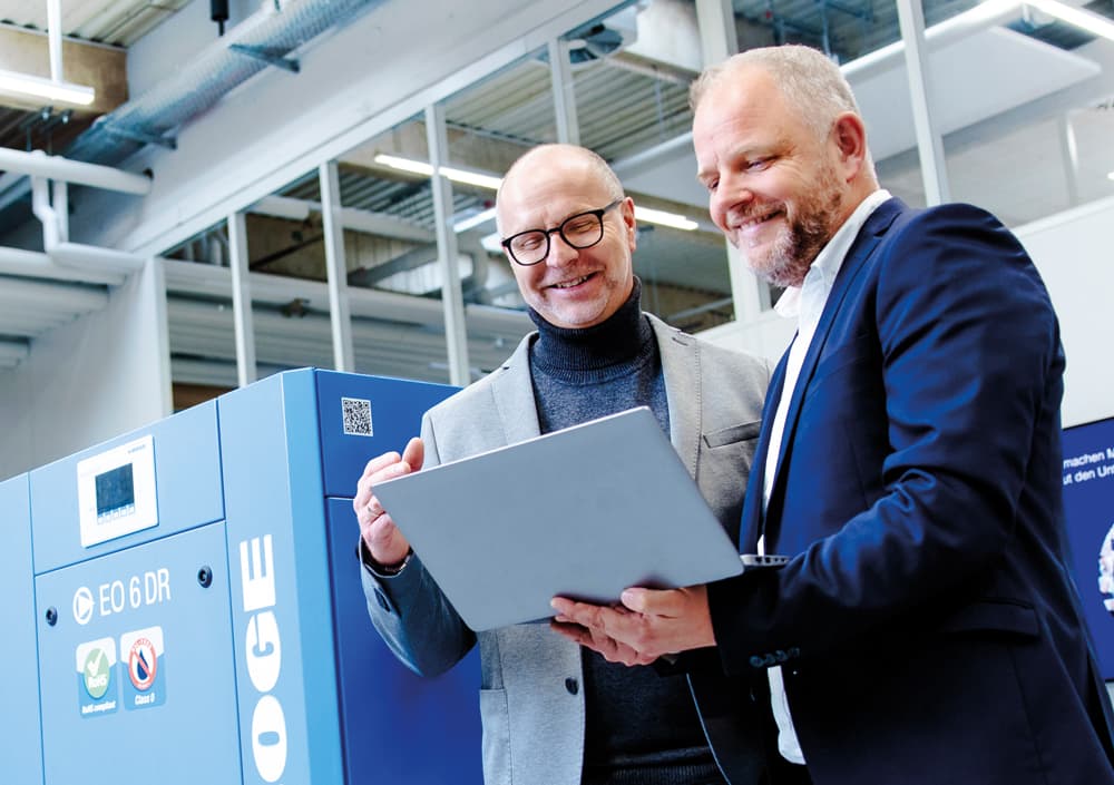 Zwei Maenner im Dialog, die einen Laptop festhalten und vor einem Kompressor stehen