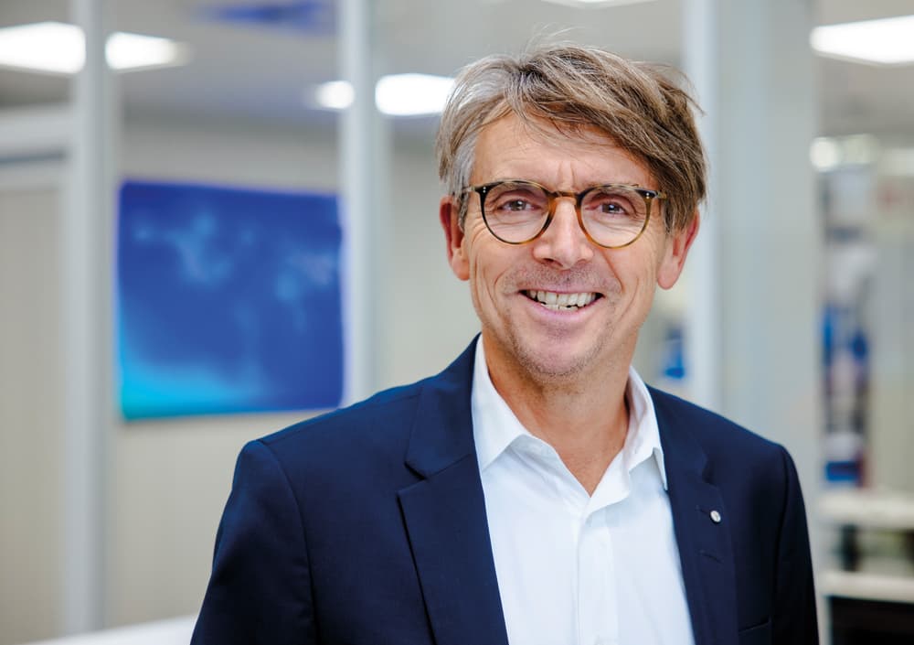 BOGE Marketing Mitarbeiter in dunkelblauem Anzug, Brille und weißem Hemd in einem Buero laechelt in die Kamera