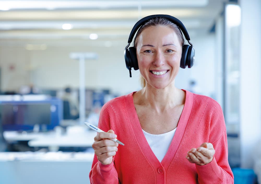 Freundliche BOGE Supportmitarbeiterin in roter Strickjacke mit Headset und Stift in der Hand in einer Büroumgebung