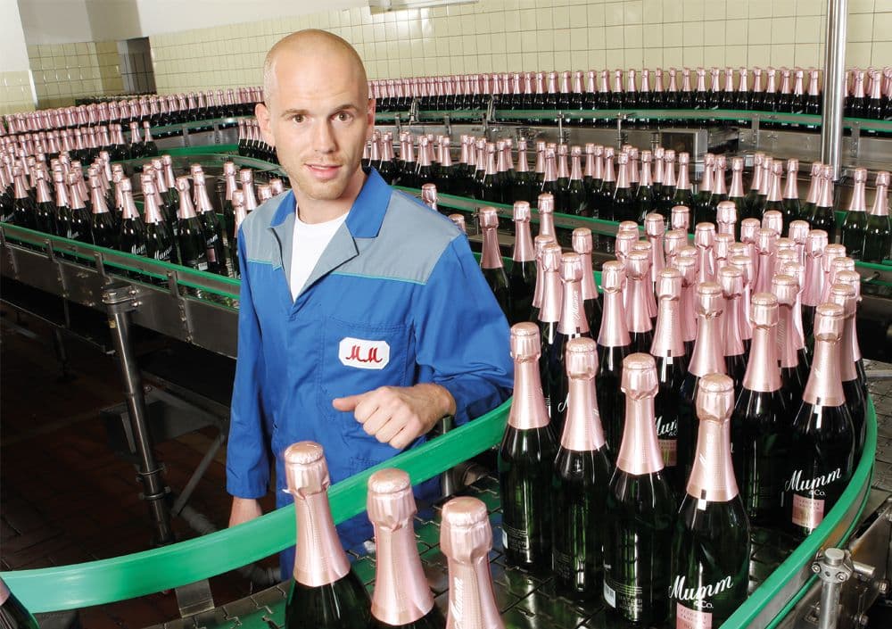 Fließband mit Sektflaschen der Marke MUMM, in dessen Mitte ein Mitarbeiter in die Kamera guckt