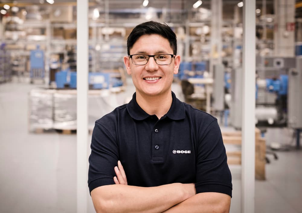 Ein lächelnder Servicetechniker von BOGE mit Brille und dunkelblauem Polohemd steht mit verschränkten Armen in einer industriellen Umgebung mit Kompressoren und Regalen