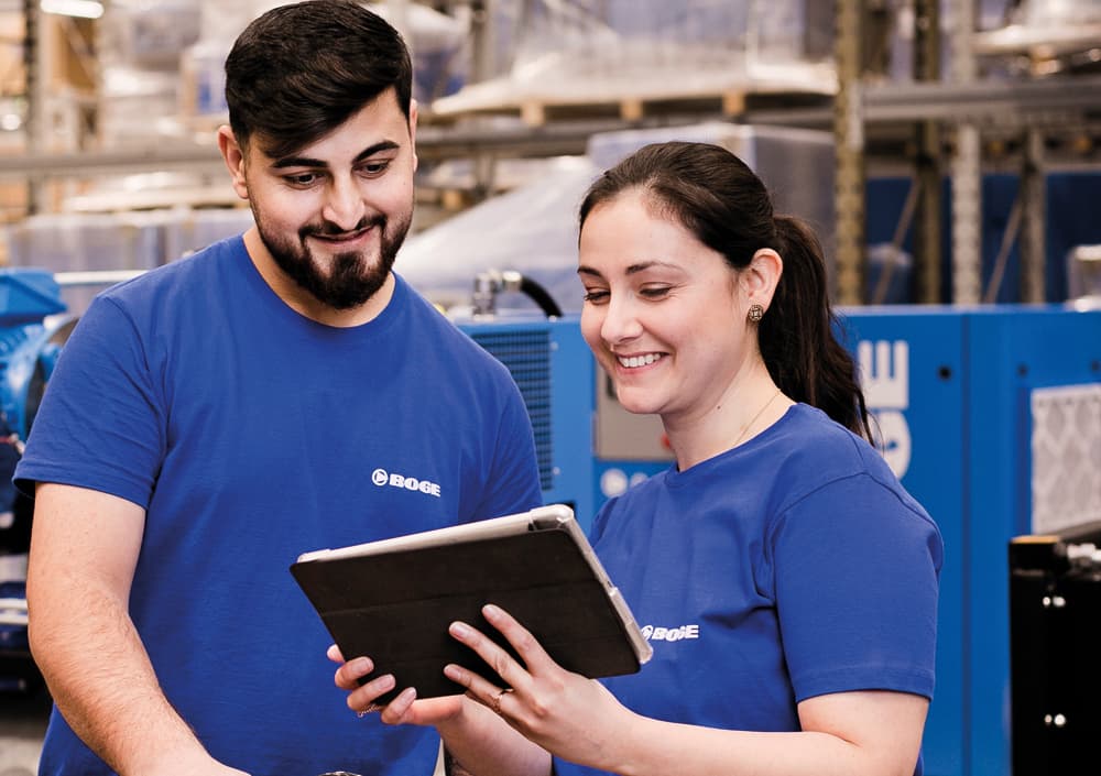 BOGE Logistikmitarbeiter stehen zu zweit in BOGE Kleidung in der Werkshalle, die Mitarbeiterin hält ein Tablet in der Hand und zeigt dem zweiten Mitarbeiter etwas