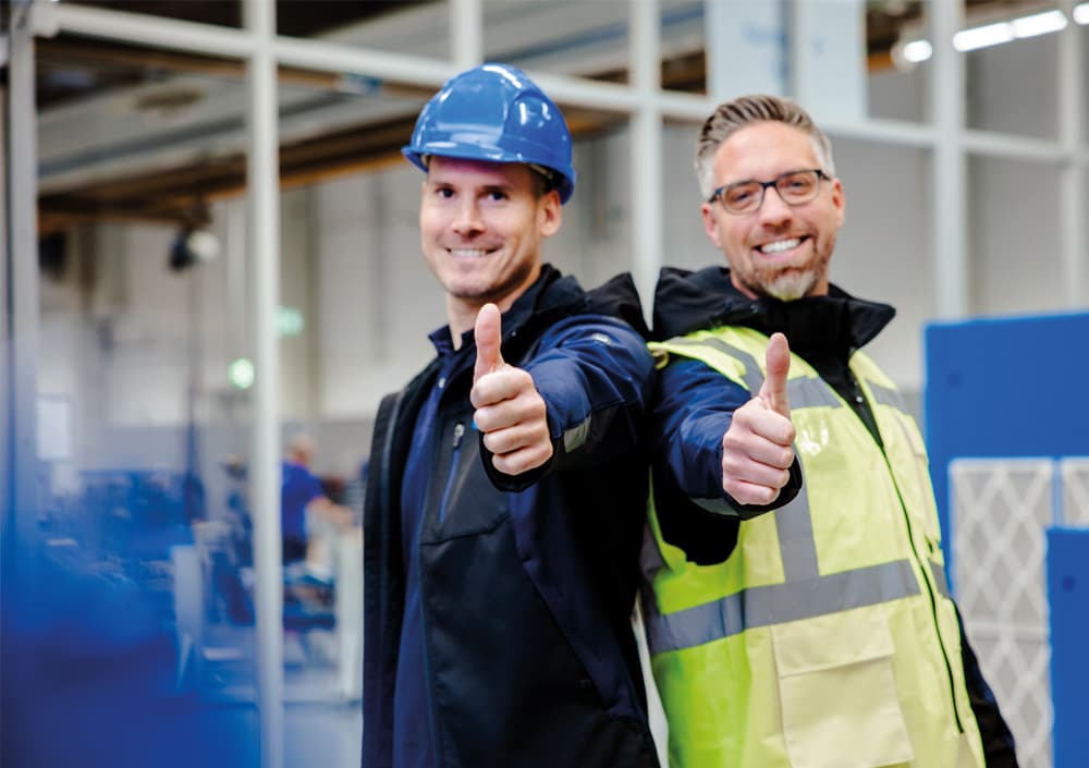 Zwei BOGE Servictechniker mit blauem Helm in einer Produktionshalle bei BOGE strecken laechelnd den Daumen nach oben