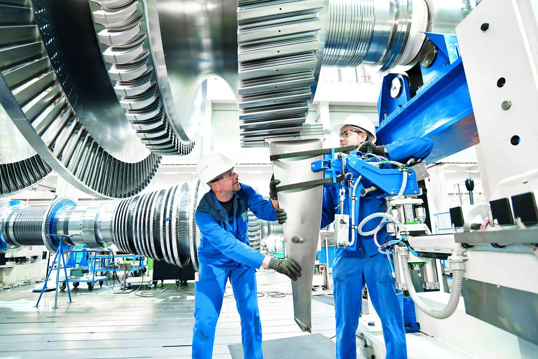 Two engineers in blue uniforms and helmets work on assembling a large turbine blade in an industrial setting.