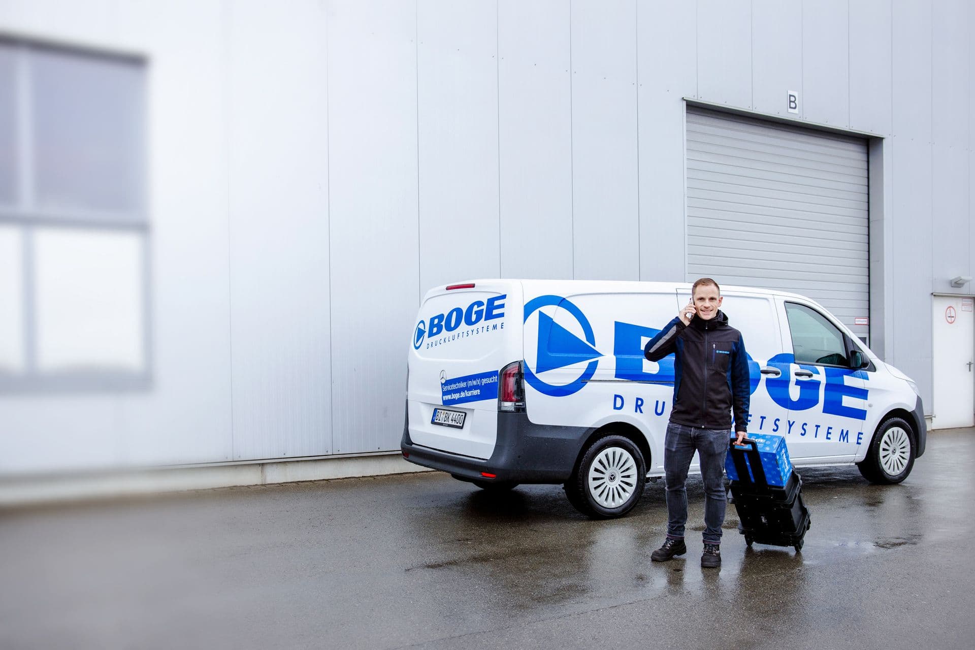 BOGE Kundendienst Mitarbeiter steht telefonierend mit einem Koffer und einem BOGE Paket vor einem BOGE Service-Fahrzeug