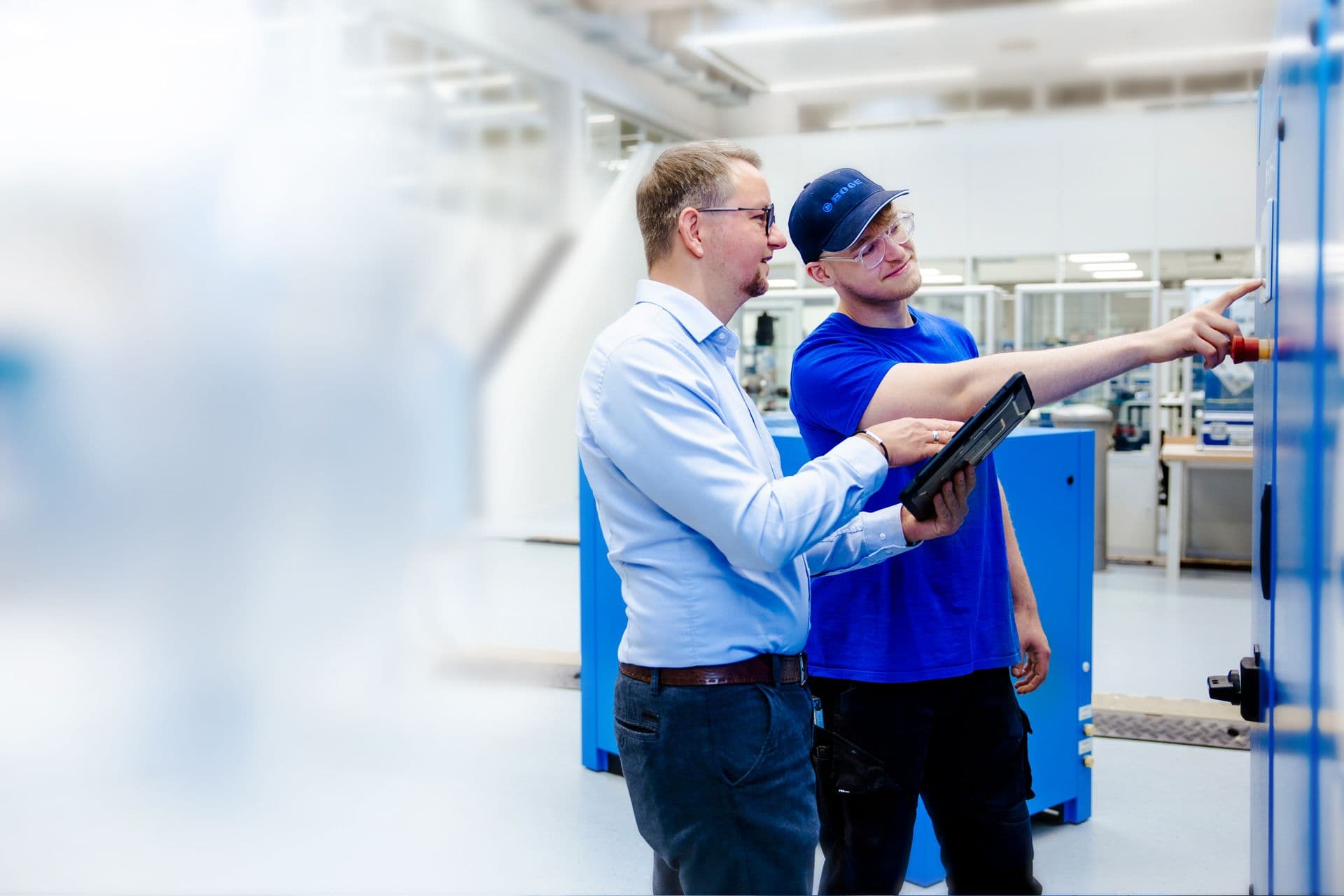 BOGE Servicetechniker in blauem T-Shirt in einer hellen Werkshalle deutet auf Steuerung eines Kompressors im Dialog mit Kunde am Tablet