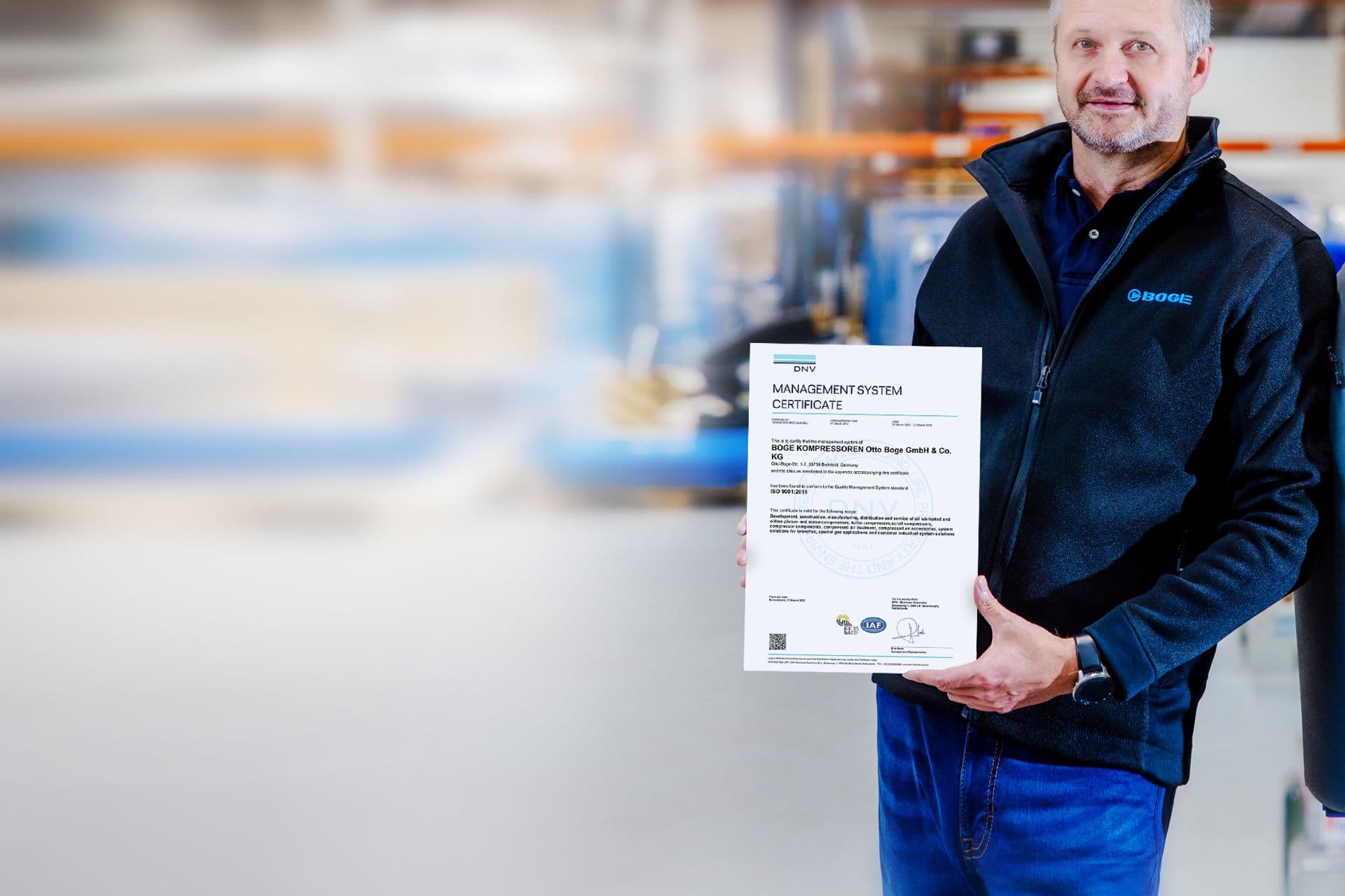 BOGE Mitarbeiter lehnt an einen Kompressor und hält das Management System Certificate in der Hand