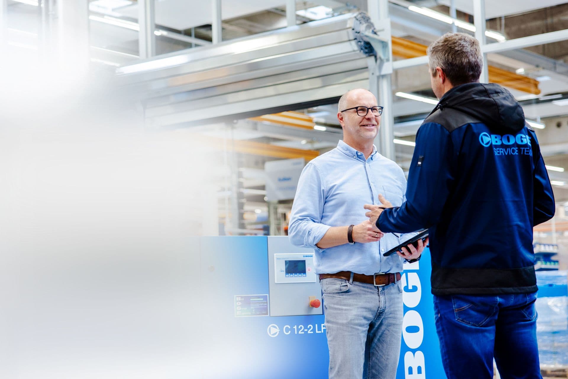 BOGE Servictechniker im Gespraech mit einem Kunden neben einem Schraubenkompressor in einer Fabrikhalle