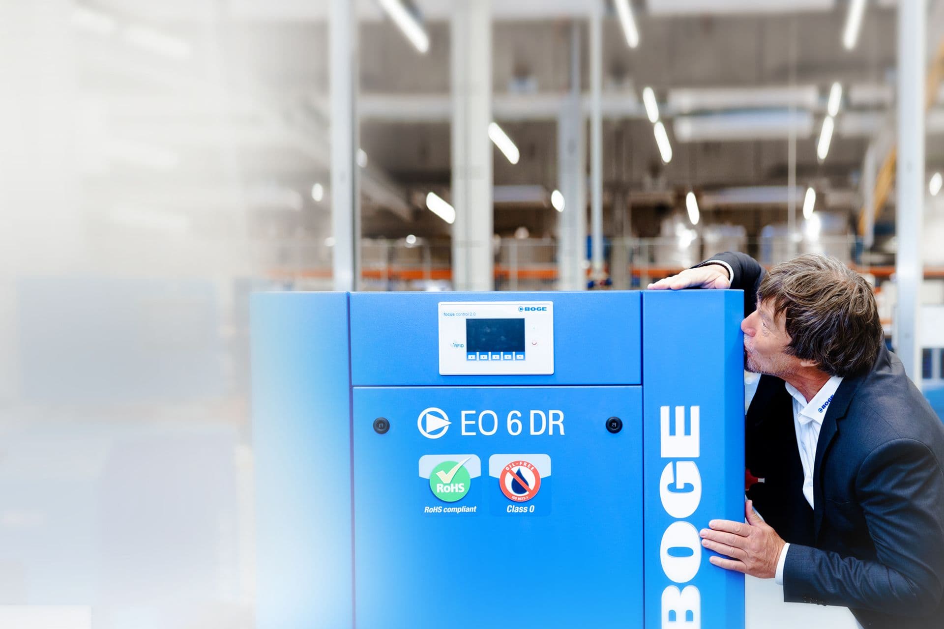 Ein BOGE Mitarbeiter in Anzug kuesst einen blauen Scrollkompressor in einer Fertigungshalle
