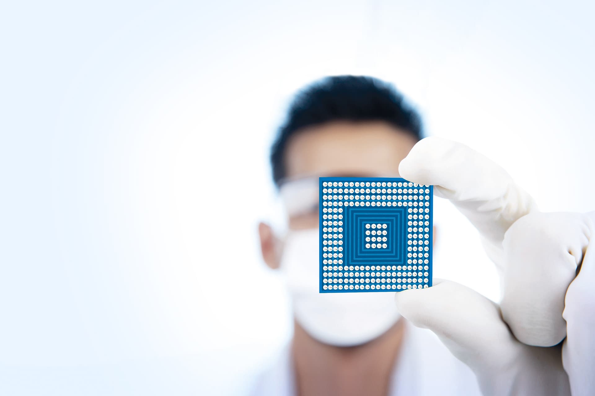 Ein Mann mit Mundschutz und weißen Einweghandschuhen hält ein elektronisches Bauteil, einen Chip in den Vordergrund.