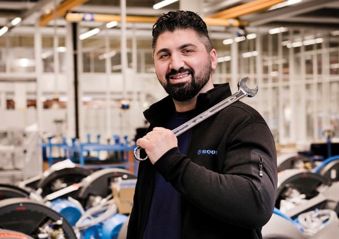 Mechaniker vor einer Vielzahl an Kompressoren in Produktionshalle guckt freundlich in die Kamera